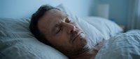 Middle-aged man with dark hair and facial stubble sleeping peacefully on white pillows in bed during daytime, suitable for wellness and health content.