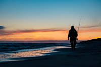 Silhouette of lone fisherman walking along beach at sunset with fishing rod, dramatic orange and blue sky reflects on wet sand, peaceful coastal scene.