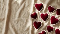 Handmade red felt heart shapes with decorative stitching scattered on natural linen fabric background for Valentine's Day crafts and romantic decorations.