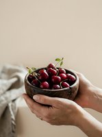 Fresh dark red cherries with stems in a wooden bowl held by hands against neutral background, suitable for food photography, healthy eating, organic produce, and culinary content.