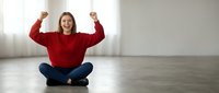 Young Caucasian woman in red sweater celebrating success with raised fists while sitting cross-legged on wooden floor in bright modern interior.