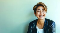Young Asian woman with short blonde hair wearing leather jacket and white t-shirt smiling genuinely against mint green wall. Casual modern lifestyle portrait.