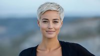Young woman with short platinum blonde pixie haircut and natural makeup smiling confidently outdoors against blurred mountain landscape background.