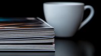 Stack of magazines with white ceramic coffee cup on black reflective surface, creating minimalist composition with dramatic lighting and selective focus.