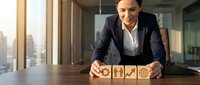 Middle-aged Caucasian female professional in dark blazer and white shirt arranging wooden blocks with business icons at office desk with city skyline view, representing strategy and corporate planning.