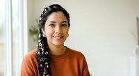 Young woman with dark braided hair and South Asian ethnicity wearing rust-orange shirt, smiling at camera in bright indoor setting with natural light and blurred background.
