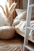 Cozy bedroom corner with pampas grass in vase, knitted pouf, jute rug, and rumpled white bedding creating warm neutral boho atmosphere for interior design.