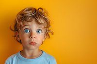 Surprised young Caucasian child with bright blue eyes and curly blonde hair wearing light blue t-shirt against vibrant yellow background, expressing wonder and amazement.