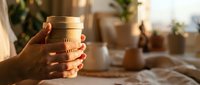 Woman hands holding warm coffee cup in cozy morning sunlight with plants and soft home interior background for relaxation and comfort lifestyle.