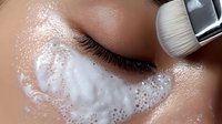 Close-up of facial cleansing process with white foam cleanser being applied under eye area using soft makeup brush, demonstrating skincare routine and beauty treatment.