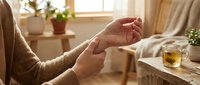 Person massaging wrist at home, cozy interior with herbal tea and plants, concept of joint pain relief and self-care wellness routine.
