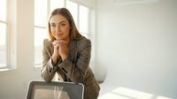 Professional young Caucasian businesswoman in gray suit sitting at desk with laptop in bright modern office, smiling confidently at camera.