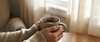 Woman hands holding warm coffee mug in cozy morning light by window. Relaxing home atmosphere with soft natural lighting and comfortable sweater sleeves.
