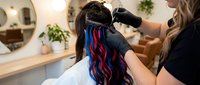 Hairstylist applying vibrant blue and red hair extensions to client with long dark hair in modern salon setting with mirror and plants.