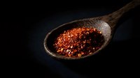 Red chili pepper flakes in rustic wooden spoon on dark background for culinary cooking and spice seasoning applications.