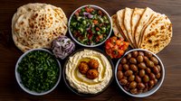Middle Eastern appetizer spread with hummus, falafel, fresh herbs, olives, pita bread and traditional mezze dishes served on wooden table background.