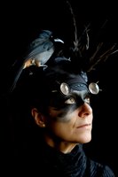 Mysterious woman in dark steampunk costume with bird headdress and silver goggles against black background, dramatic side lighting creates theatrical mood.