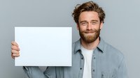 Caucasian man with beard and wavy hair holding blank white sign against gray background, smiling confidently while displaying empty poster for advertising message.