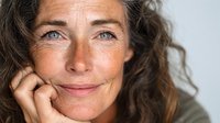 Mature woman with natural beauty, curly hair and blue eyes smiling gently against neutral background. Authentic portrait showing confidence and wisdom of aging gracefully.