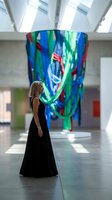 Elegant blonde woman in black evening gown admiring colorful contemporary art installation in modern gallery space with natural lighting.