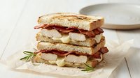 Gourmet turkey club sandwich with crispy bacon and melted cheese on toasted artisan bread, garnished with fresh rosemary on parchment paper.