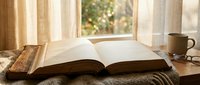 Open book on wooden table by sunny window with coffee mug creating cozy reading atmosphere for relaxation and learning in warm natural light.