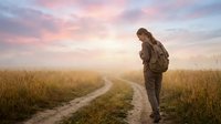 Young adult male hiker with backpack standing on rural dirt path through golden wheat field at sunrise, looking down contemplatively during outdoor adventure journey.