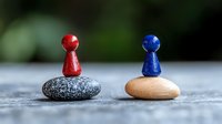 Red and blue game pieces balanced on contrasting smooth stones against blurred background, representing balance, opposition, and strategic decision making.