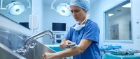 Male surgeon washing hands at sink in modern operating room with surgical lights and medical equipment in sterile hospital environment.