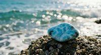 Turquoise gemstone resting on rocky shore with sparkling ocean waves and sunlight reflections creating peaceful coastal meditation scene for wellness content.