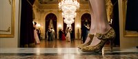 Elegant golden embroidered high heel shoe in a luxurious ballroom with chandeliers and dancing couples in the blurred background.