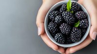 Fresh ripe blackberries in white ceramic bowl with mint leaves held in female hands against gray background, top view with copy space.