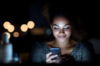 Young African American woman with curly hair smiling while using smartphone in dark room with bokeh lights, face illuminated by phone screen glow.