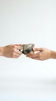 Two hands holding a rustic handmade ceramic mug together on a light background, symbolizing sharing, connection, and mindful living.