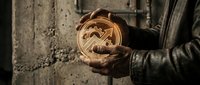 Craftsperson holding wooden circular ornament with intricate carved geometric pattern against weathered concrete wall, showcasing traditional woodworking artistry and handmade craftsmanship.
