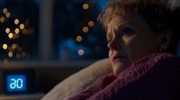 Mature woman with short blonde hair lying awake at night, digital alarm clock showing 2:00 AM. Dark room with bokeh lights in background suggests insomnia or sleep issues.