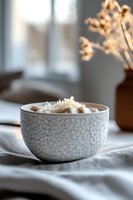 Creamy hot chocolate topped with coconut flakes served in textured white ceramic bowl against soft window light, creating cozy morning atmosphere.