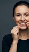 Middle-aged woman with dark hair and fair complexion smiling while flossing teeth against neutral background. Dental hygiene and oral care concept.