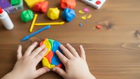 Child hands playing with colorful modeling clay on wooden table surrounded by art supplies and craft materials for creative learning activities.