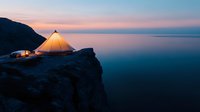 Illuminated camping tent glowing on rocky cliff edge at sunset, overlooking calm ocean with pink and blue sky, creating atmospheric outdoor adventure scene.