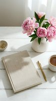 Pink peonies in white vase on rustic desk with linen notebook, gold pen, tea cup, and paper clips in soft natural light.