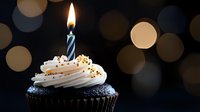 Single chocolate cupcake with white frosting and lit blue striped birthday candle on dark background with soft bokeh lights creating festive atmosphere.