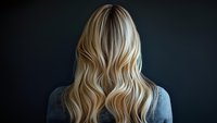 Back view of woman with long blonde wavy hair against dark background, showcasing modern hairstyle with highlights and natural texture.
