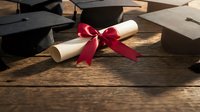 Graduation diploma with red ribbon bow surrounded by mortarboard caps on rustic wooden table, celebrating academic achievement.