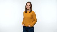 Young woman with long brown hair and fair complexion wearing mustard yellow ribbed sweater and dark jeans, smiling at camera against white background for portrait use.