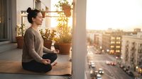 Woman in casual sweater sitting on balcony at sunset, holding cup, overlooking urban cityscape with golden hour lighting and potted plants for relaxation and lifestyle content.