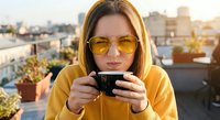 Young woman in yellow hoodie and sunglasses enjoying hot beverage on urban rooftop terrace during golden hour sunset with city skyline background.