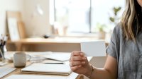 Young woman with medium skin tone holding blank white card at desk, wearing casual gray shirt, suitable for business communication, marketing, and corporate messaging concepts.