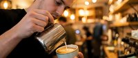 Professional barista creating latte art in coffee cup at busy cafe, pouring steamed milk from metal pitcher into espresso drink during morning service.