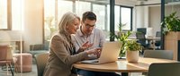 Mature woman and middle-aged man collaborating on laptop at wooden desk in bright modern office with plants and natural light, discussing work project together.